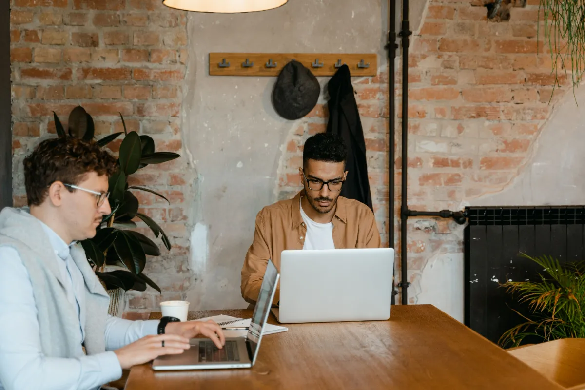Two men working on laptops at a shared wooden table in a brick-walled café